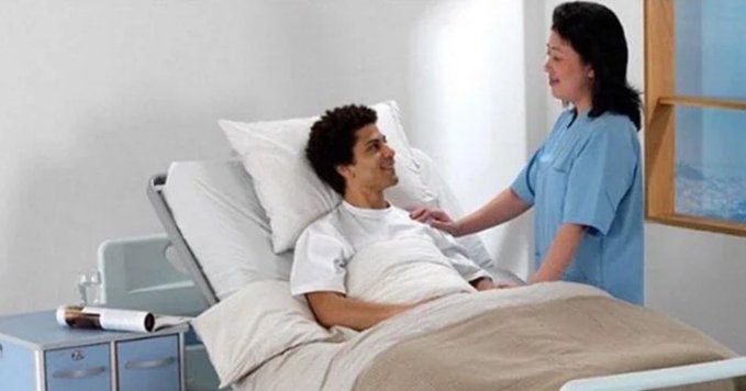 A man in a hospital bed smiling at a female caregiver in blue scrubs who is standing beside his adjustable bed in a well-lit room.
