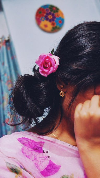 A close-up of a woman from behind with a pink rose tucked in her dark hair bun, wearing dangling earrings and a pink floral top, with a colorful floral decoration on the wall behind her.