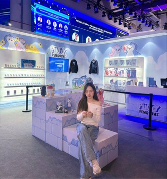 A young woman sitting on a block-patterned bench inside a brightly lit 'PENGUINS' retail store, surrounded by shelves of merchandise and a display of clothing.