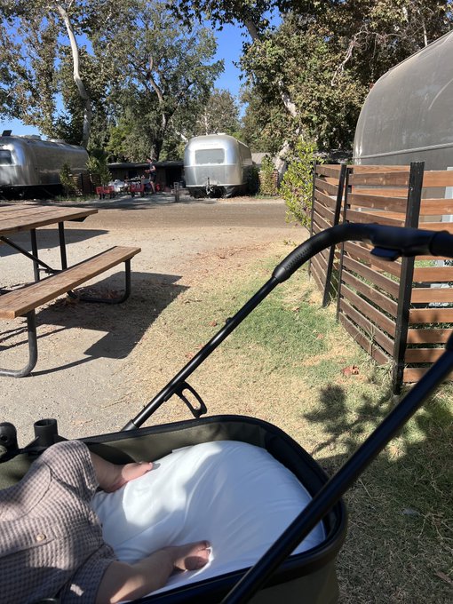 A baby in a white outfit sitting in a stroller at an outdoor campground featuring shiny silver Airstream trailers, a wooden picnic table, and surrounding trees.