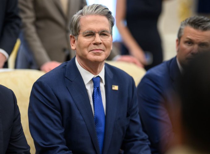 A portrait of Scott Bessent, U.S. Treasury Secretary, wearing a navy blue suit and blue tie, seated in what appears to be a formal meeting room with other officials.