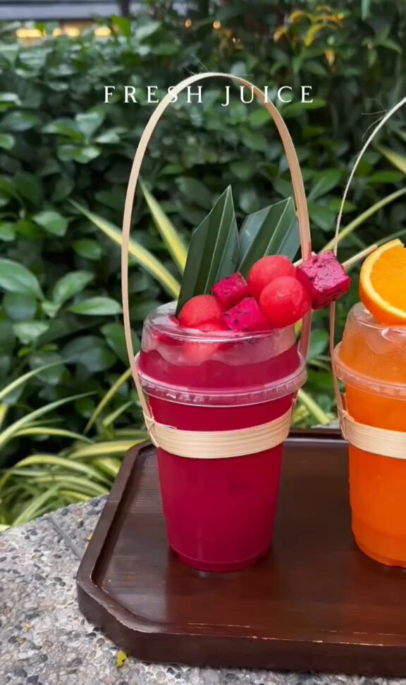 The image features two cups of juice, one red and one orange, placed on a wooden tray or table. The cups are filled with fruit juices, possibly watermelon and orange juice. A straw can be seen inside each cup. The background includes green plants that add a natural touch to the scene. The post is titled "Fresh Juice" and has a caption about the drinks being served.