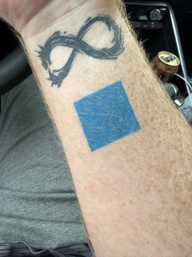 A forearm tattoo featuring a thorned thorn crown/infinity design above a blue square, with a cigarette lighter visible in the background.