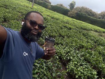 A man with a beard and sunglasses takes a selfie in a lush green tea field while holding a takeaway coffee cup and wearing a gray t-shirt.