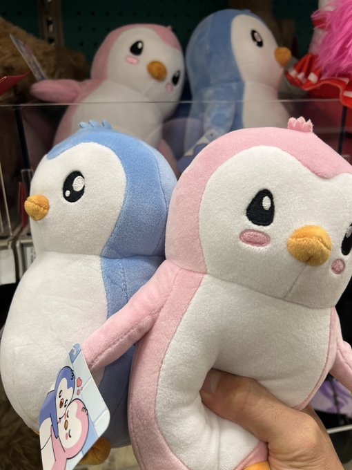 A hand holds a soft pink penguin plush toy in front of a store display showing a similar blue penguin plush and other stuffed animals.