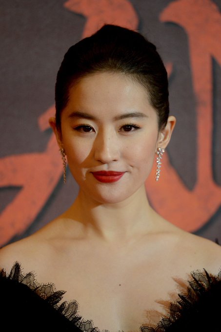 A close-up portrait of an Asian woman with her hair pulled back, wearing bold red lipstick, dangling earrings, and a black dress with a lace neckline, photographed against a dark background with red lettering.