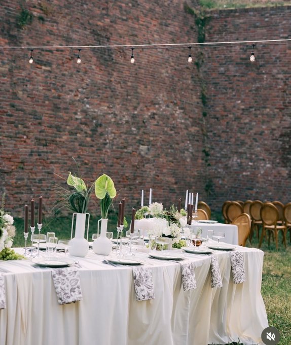 An elegant outdoor wedding reception setup with a long table draped in white linens, decorated with white flowers, candles, and white vases, set against a rustic brick wall with string lights overhead.