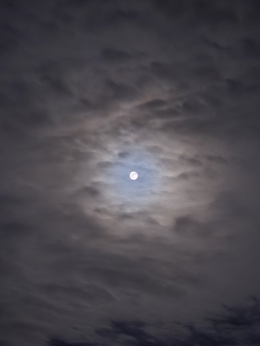 A full moon shines brightly through a break in dark, wispy clouds against a night sky.