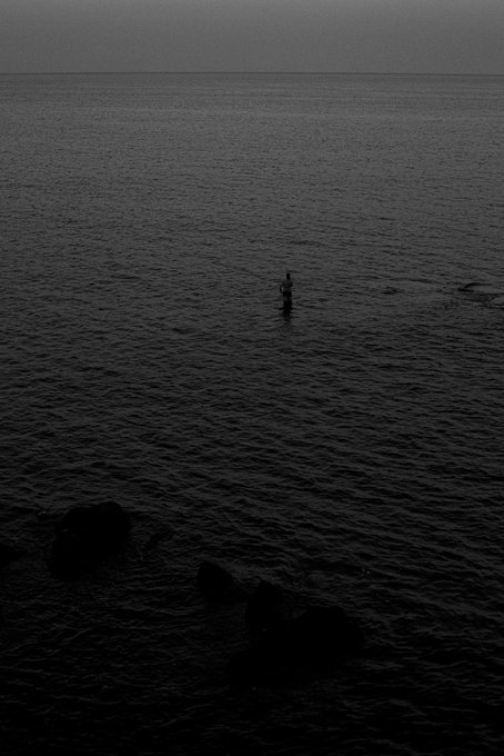 A dark, moody photograph of a lone figure standing waist-deep in calm ocean water at dusk, with rocks visible in the foreground.