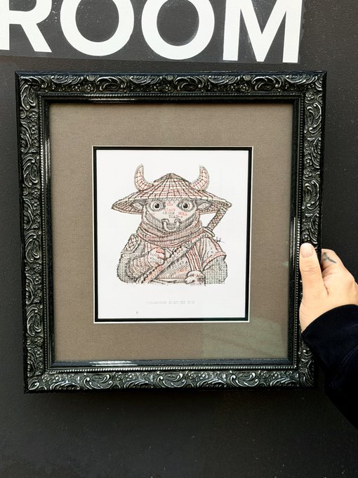 A framed piece of art featuring an intricate cross-stitch or embroidered design of a horned, masked creature wearing a samurai-style hat, held by a hand with a tattoo visible on the wrist against a black wall with partial white 'ROOM' text.