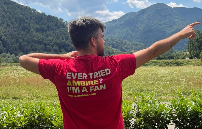 A man from behind wearing a red t-shirt that reads 'EVER TRIED AMBIRE? I'M A FAN www.ambire.com', pointing toward a scenic mountain landscape.