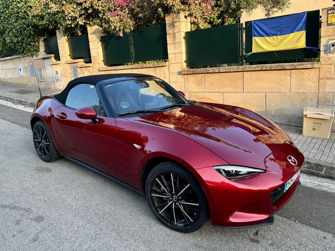 Red Mazda MX-5 convertible roadster parked on a street in front of a stone wall with a Ukrainian flag and green shutters.