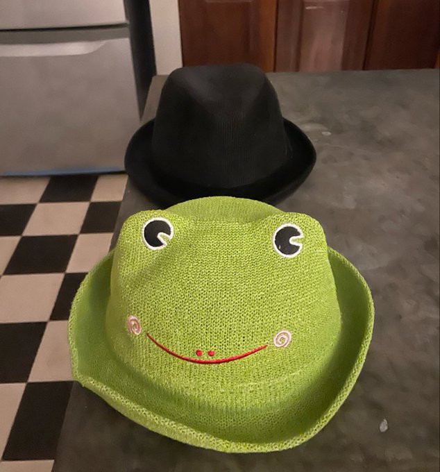 Two hats placed on a concrete surface over a checkered floor - a black fedora in the background and a bright green crocheted frog bucket hat with eyes and a smiling face in the foreground.