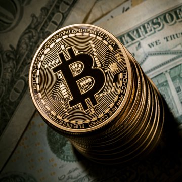 A stack of physical Bitcoin tokens rests atop U.S. currency, with the inscriptions "BITCOIN DIGITAL DECENTRALIZED PEER TO PEER" and "VIRES IN NUMERIS FINE COPPER" clearly visible around the coin's edge, visually reinforcing the article's focus on Bitcoin's price movement amid macroeconomic uncertainty.