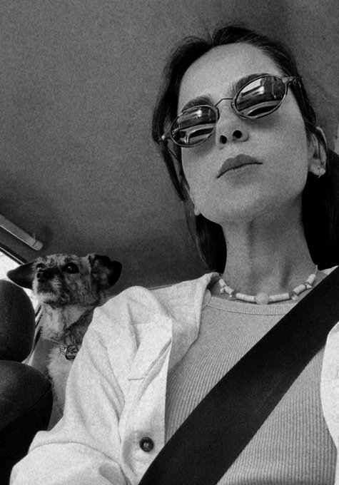 A black and white selfie of a woman wearing round sunglasses and a beaded necklace, seated in a car with her seatbelt fastened, while a small dog looks at the camera from the back seat.