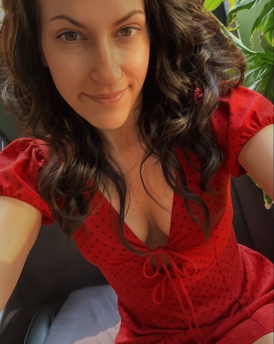 The image shows a smiling woman taking a selfie in a red polka dot dress with a tie-front detail, posing indoors near a green plant. The caption plays into her post by inviting followers—especially "Reply Guys"—to DM her if they unfollow, with the cheerful, self-assured photo adding a playful, unbothered confidence to the request for honest feedback.