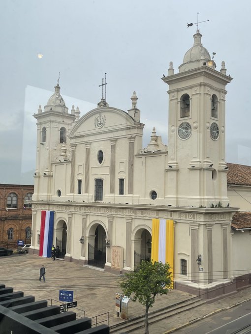 A large white cathedral with twin bell towers and domes stands in a plaza, decorated with red, white, and blue banners as well as a yellow banner.