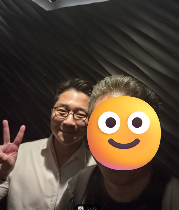 A selfie of two people, with one person wearing glasses and a white shirt making a peace sign, while the other person's face is covered by a yellow smiley emoji sticker.
