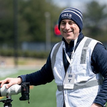 A photograph of a smiling man wearing a blue 'VALE' beanie and a reflective safety vest with a lanyard and ID badge, standing outdoors with his hand resting on equipment, possibly a camera rig or spotting scope.