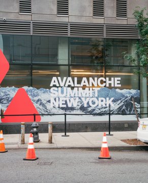A street view shows the entrance to the Avalanche Summit NYC event, with a large banner displaying "AVALANCHE SUMMIT NEW YORK" against a mountain backdrop, accompanied by orange traffic cones and barriers on the sidewalk outside the venue. The image depicts the setup for Day 2 of the summit, which is hosting speakers including MoonPay, Live Nation, Sports Illustrated, the Cleveland Cavaliers, Kevin O'Leary, FIFA, Dinari, and Kalshi.