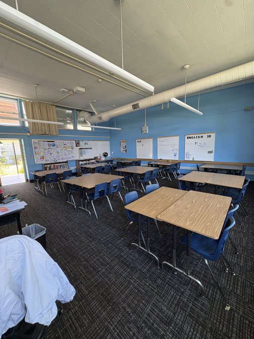 An empty classroom features rows of blue chairs pushed into wooden desks, with 'ENGLISH 10' clearly written on a whiteboard at the front.