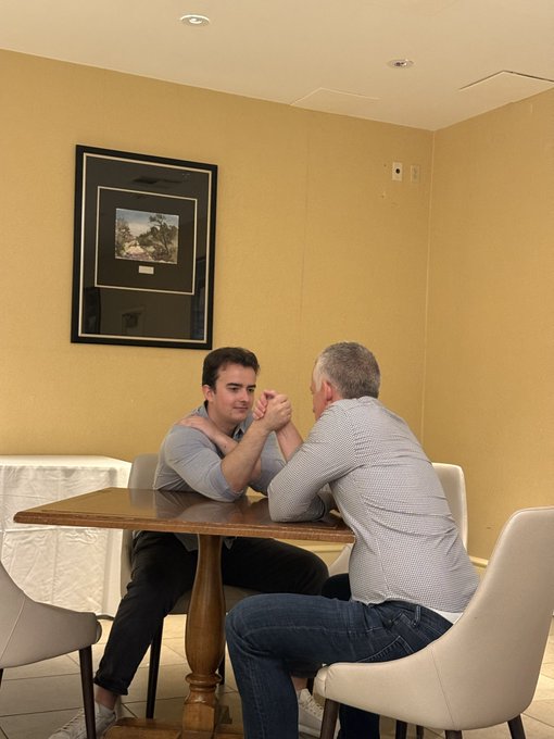 Two men sit across from each other at a small wooden table, engaged in an arm wrestling match in a simple room with yellow walls and a framed picture.