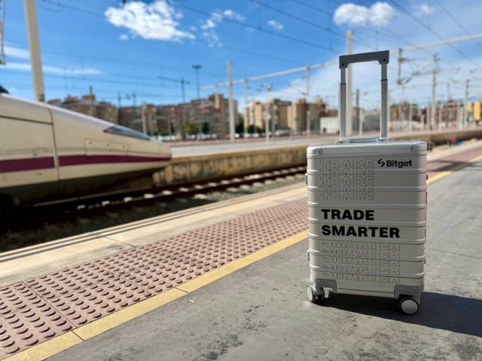 A silver Bitget-branded suitcase labeled "TRADE SMARTER" sits on a sunny train platform, ready for departure. Looks like someone's packing up to take their crypto trading on the go—Gm, Africa! Ready to ride the markets with you! 🚄💼
