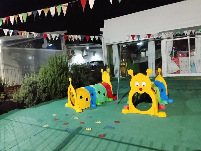 A nighttime outdoor scene showing colorful caterpillar-shaped children's play tunnels on a green surface in front of a house decorated with festive flag bunting overhead.