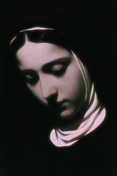 A dark, dramatic close-up portrait of a woman in a nun's habit with her head bowed in shadow.