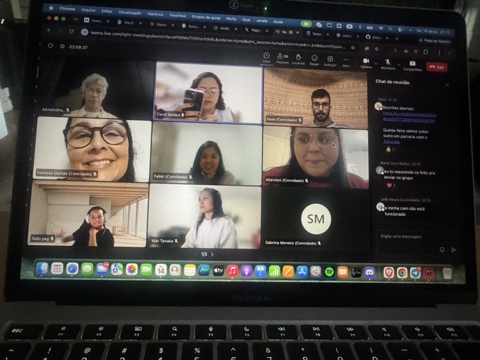 A MacBook Air laptop displays a Teams video meeting in progress with nine participant tiles visible, including names like 'Vanessa Santos', 'Carol Santos', 'Ivan (Convidados)', 'Fabbi', 'Marcelly', and chat messages in Portuguese on the right side.