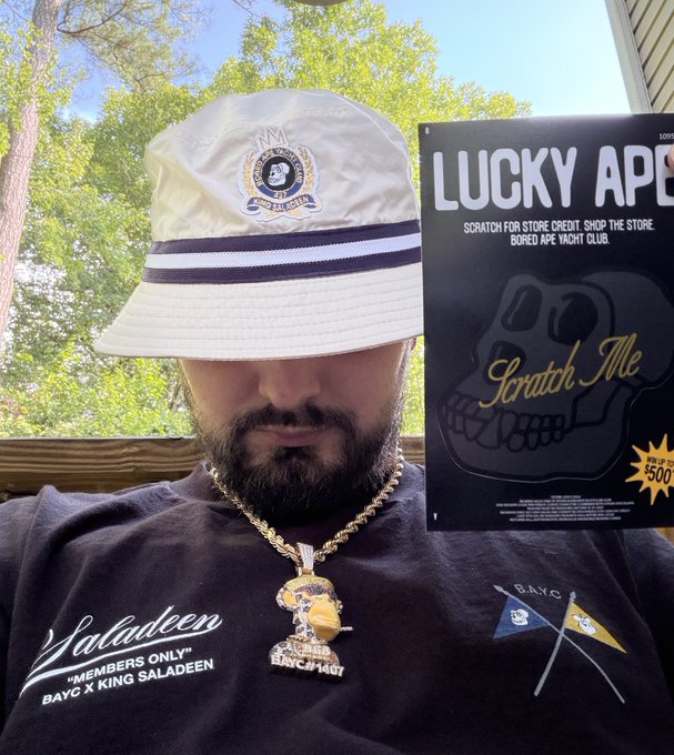 A bearded man wearing a white bucket hat and a black Saladeen 'BAYC x King Saladeen' t-shirt, holding a 'Lucky Ape' Bored Ape Yacht Club scratch-off card.