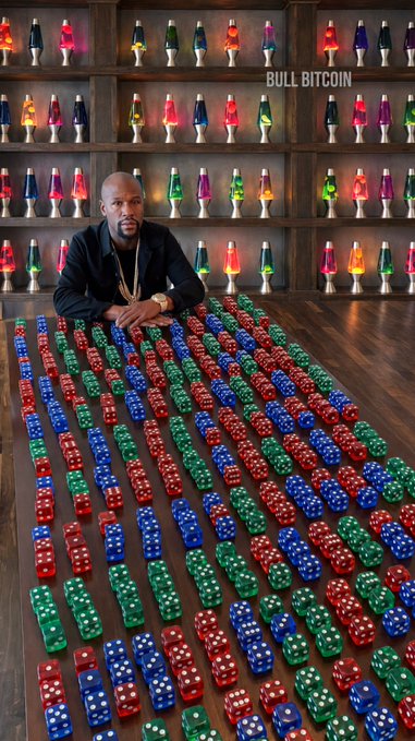 A man sits at a table covered with rows of dice arranged in bullish patterns (red, green, and blue), backed by shelves of glowing lava lamps, with "BULL BITCOIN" text visible, visually representing a strong bullish sentiment among Bitcoin holders.