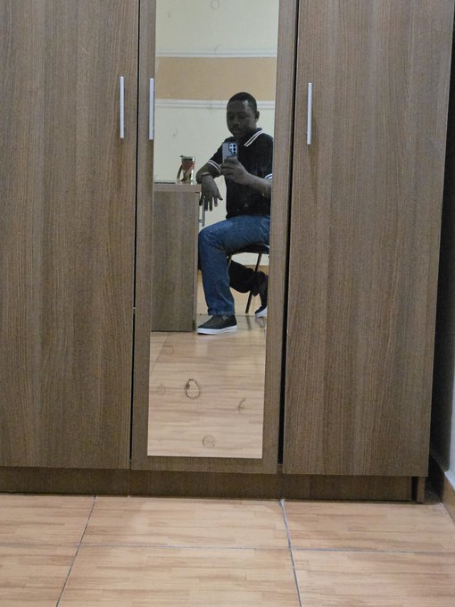 A person takes a mirror selfie with their phone while sitting on a chair in front of a wooden wardrobe, capturing a casual weekend moment that fits the "GM" (Good Morning) post about enjoying the day.