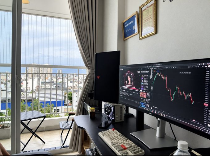 A home office setup with two monitors on a black desk, one showing a candlestick chart, with a keyboard, PC, and a view of a city from a window.