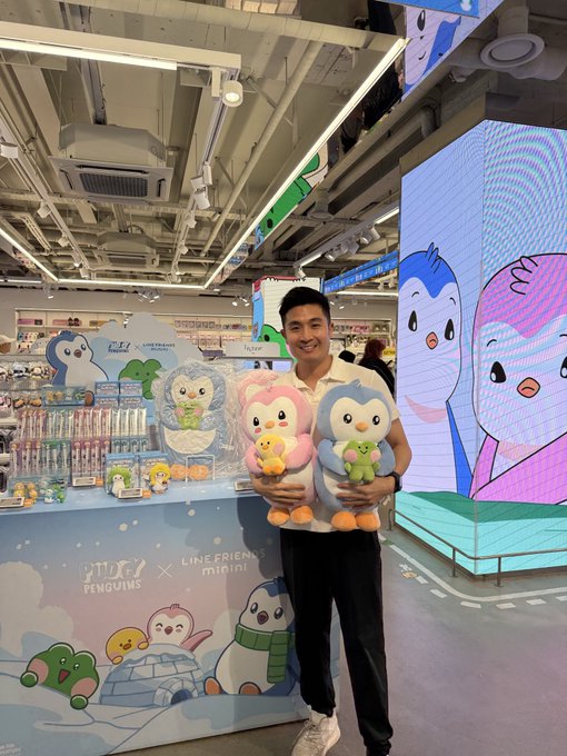 A man posing at a LINE FRIENDS minini x Snow Penguins promotional booth in a retail store, holding two penguin plush toys in front of a backdrop featuring the same characters.