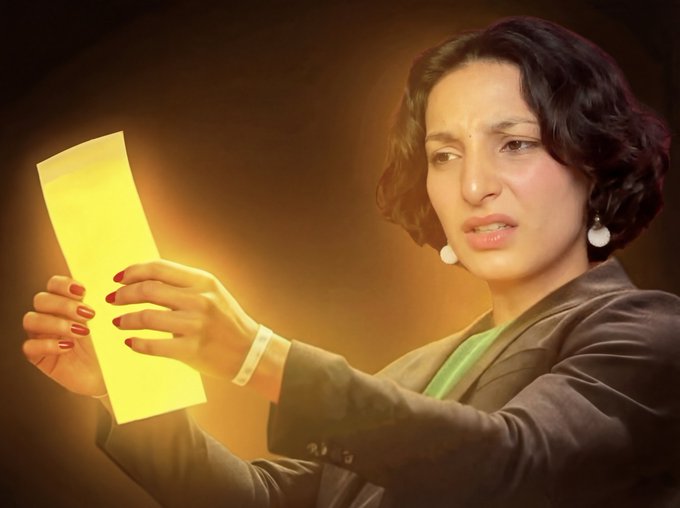 A woman with short dark hair and pearl earrings looks at a yellow piece of paper with a distressed expression.