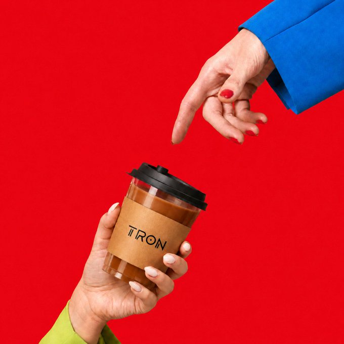 Two hands exchange a takeaway coffee cup with a cardboard sleeve labeled 'TRON' against a bright red background, one hand in a blue sleeve pointing downward while the other in a green sleeve holds the cup.