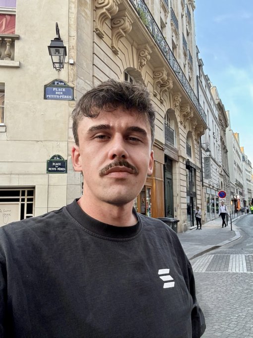 A selfie of a young man with a mustache wearing a black sweatshirt, standing on a street with Parisian-style architecture and a sign for 'Place des Petits-Pères' in the background.