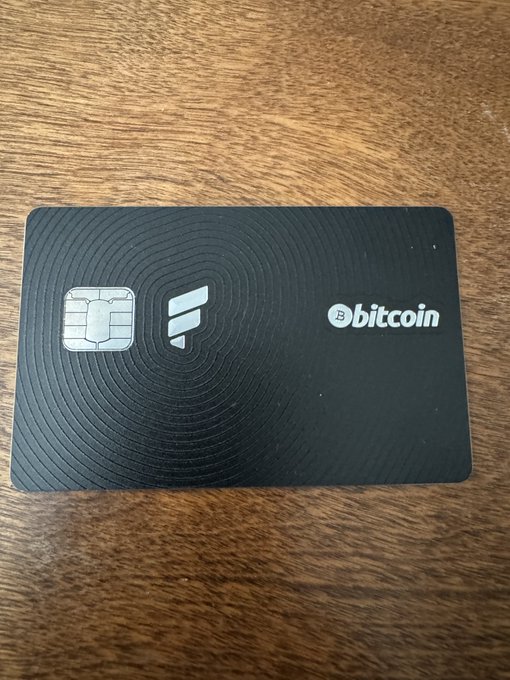 The image features a black credit card with white text, which reads "BITCOIN". The card has been used for making purchases and is now being used as an investment tool by someone who wants to earn Bitcoin. The card is placed on top of a wooden table or countertop, indicating that it was recently used or is about to be used again.