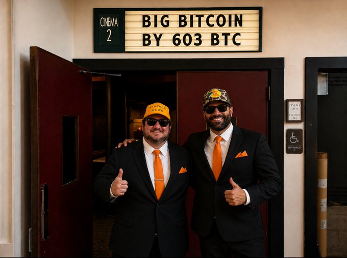 Two men in black suits with orange ties and Bitcoin hats posing in front of a cinema door marked 'CINEMA 2 BIG BITCOIN BY 603 BTC'.