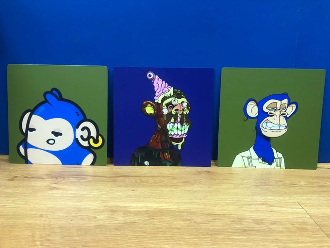 Three square metal plates lean against a blue wall on a wooden floor, featuring NFT artwork from the Bored Ape Yacht Club (BAYC), Mutant Ape Yacht Club (MAYC), and the honorary Balloons collection, with each plate displaying its respective ape character against green or blue backgrounds.