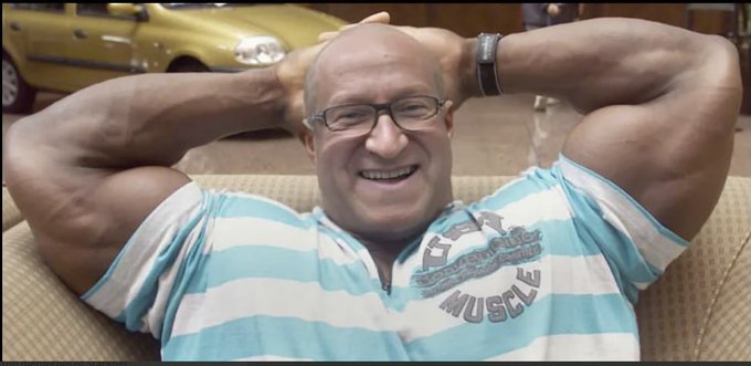 A muscular, smiling bald man with glasses wearing a blue and white striped 'MUSCLE' shirt flexes both biceps while sitting on a couch, with a yellow car visible in the background.
