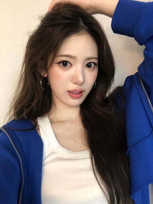 A selfie of an Asian woman with long dark wavy hair, wearing a blue zip-up hoodie over a white top and silver hoop earrings.