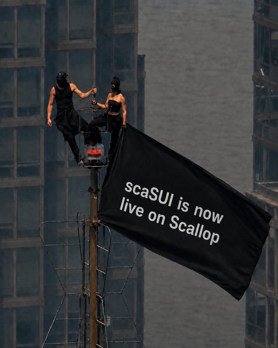 Two masked performers atop a pole displaying a black flag reading 'scaSUI is now live on Scallop' against a city skyline.