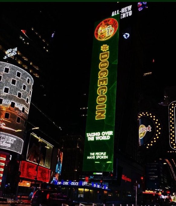 A nighttime photo of Times Square showcases a massive illuminated digital billboard prominently featuring the Dogecoin logo, a Shiba Inu dog emblem, and the slogan "#DOGECOIN - TAKING OVER THE WORLD - THE PEOPLE HAVE SPOKEN" in green and gold neon, visually reinforcing the post's bullish sentiment about the cryptocurrency's mainstream visibility.