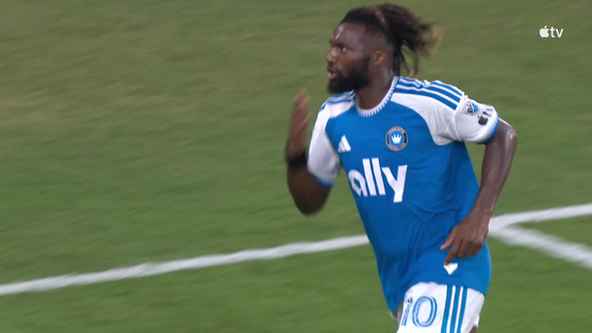 Allan Saint-Maximin, wearing a blue Charlotte FC jersey with the "ally" sponsor logo and the number 10, celebrates on the pitch after scoring his first MLS goal against DC United. The image captures his jubilant reaction as Charlotte takes a 2-1 lead.