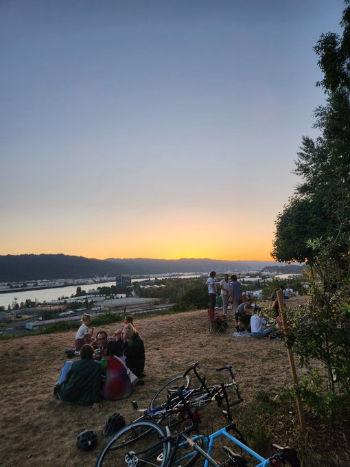 A group of people gathered on a grassy hilltop at sunset, watching the view over a river and city, with bicycles parked in the foreground.