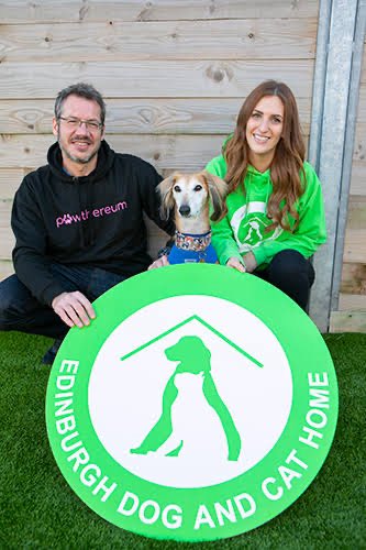 A man and woman pose with a beagle wearing a blue bandana, sitting behind a large green "Edinburgh Dog and Cat Home" sign featuring a dog and cat under a roof silhouette. The image illustrates the charity donation mentioned in the post, showing representatives (likely from Pawt erceum) celebrating their $50,000 $SAN contribution to the animal rescue organization.
