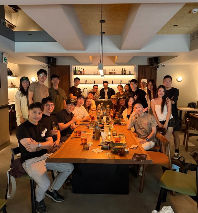 The image shows a group of people gathered around a long wooden dining table, enjoying each other's company at an event or gathering. The room has high ceilings and large windows that allow natural light to illuminate the space. There are several chairs placed around the table, indicating a social setting where guests can sit and mingle with one another.

The group consists of people from various countries, suggesting a diverse and international atmosphere. They appear to be engaged in conversation or enjoying food and drinks together. The image captures a moment of camaraderie and connection among these individuals who have come together for this special occasion.