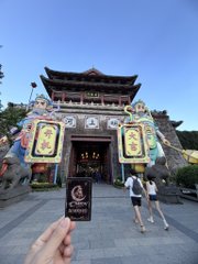 A photograph of a Chinese temple entrance with traditional architecture and guardian statues, with a hand holding a black card in the foreground.